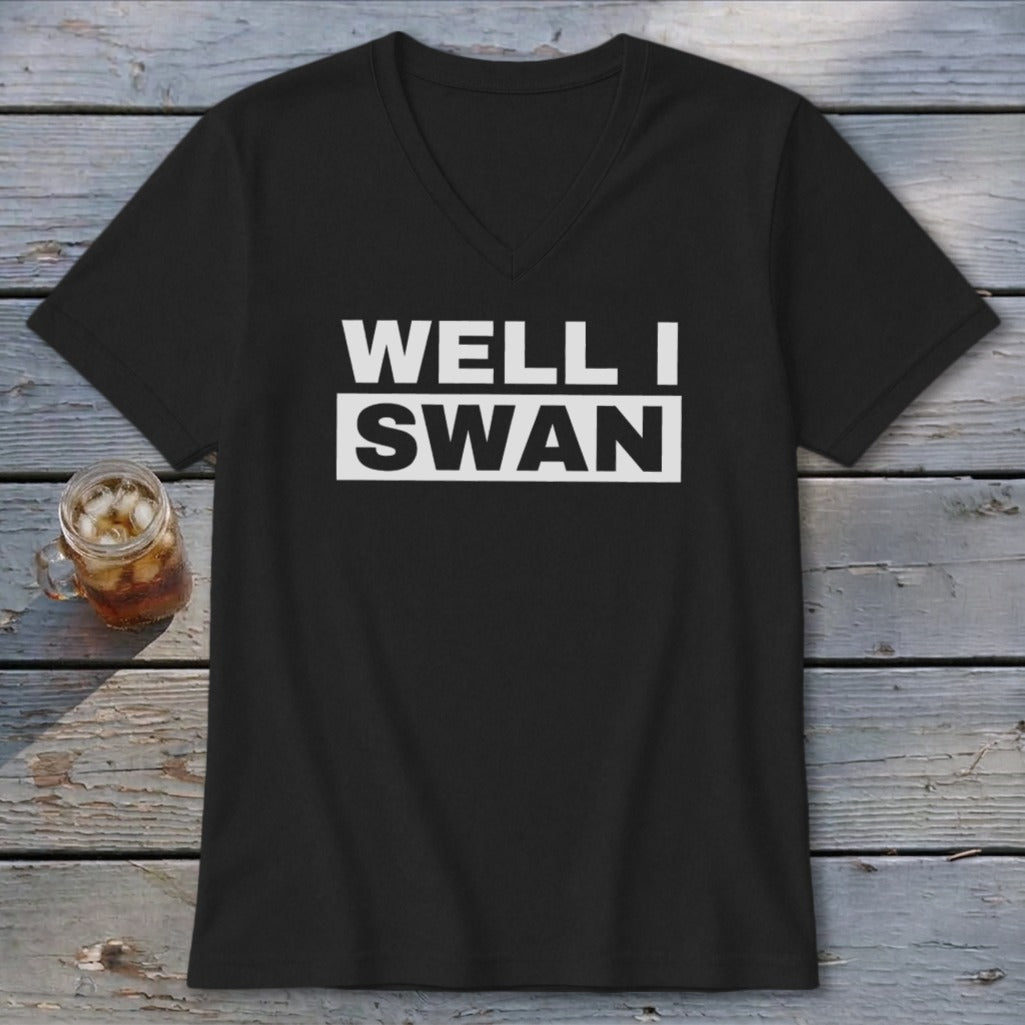 A black V-neck t-shirt with the text WELL I SWAN printed in white on the chest, laid on a wooden surface next to a glass of iced tea.