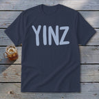 Dark blue t-shirt with light blue "YINZ" text, beside a glass of iced beverage.