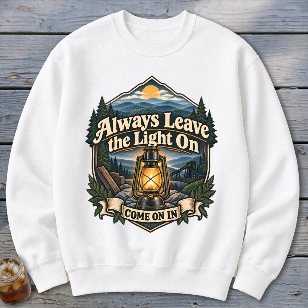 White sweatshirt with graphic of a lantern in mountain landscape with text "Always Leave the Light On COME ON IN".
