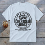A light blue t-shirt printed with a circular badge reading Certified Cornbread Judge above text about disqualification.
