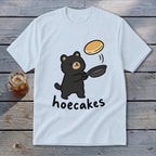 A light blue t-shirt featuring a cartoon black bear flipping a pancake in a pan, with the text hoecakes underneath.