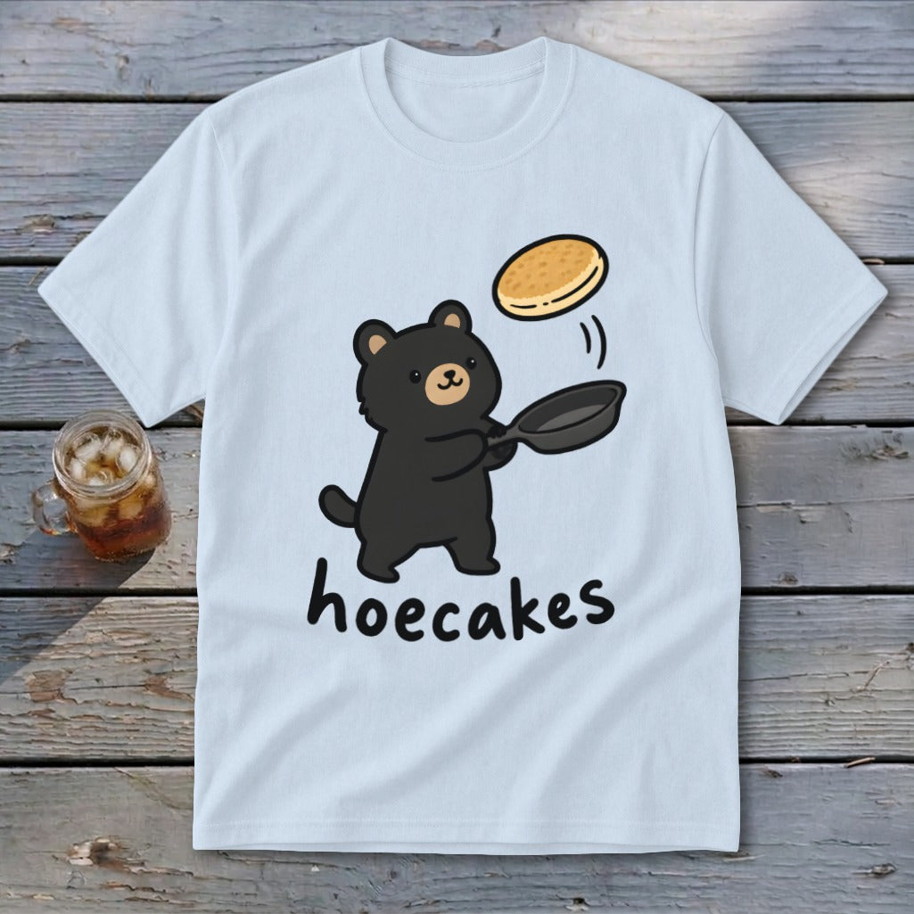 A light blue t-shirt featuring a cartoon black bear flipping a pancake in a pan, with the text hoecakes underneath.