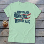 A mint green t-shirt with rolled sleeves lies flat on a rustic wooden background, featuring a vintage American flag graphic with the text MARYLAND RAISED AMERICAN MADE.