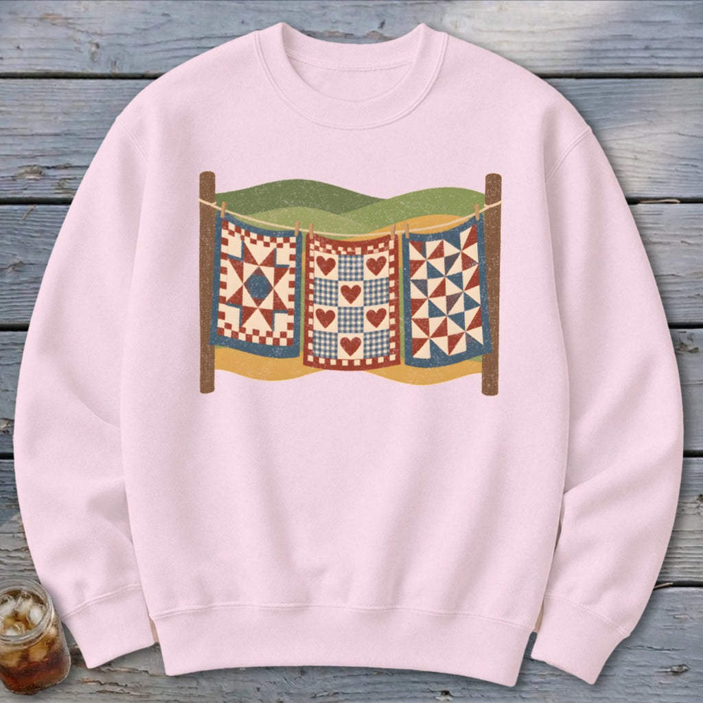 Light pink crewneck sweatshirt featuring a graphic of three colorful patterned quilts hanging on a clothesline.