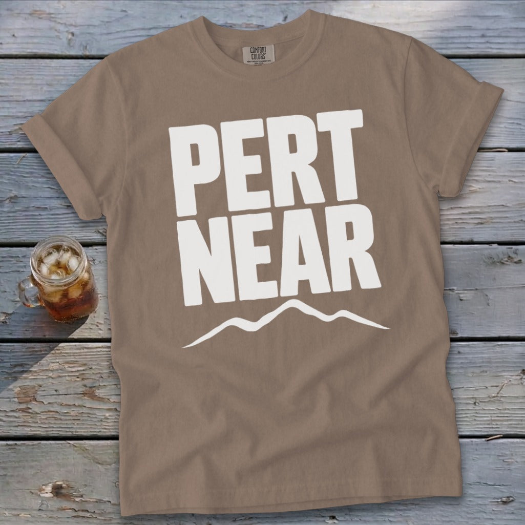 A brown crewneck t-shirt with rolled sleeves lies on a rustic wooden table, featuring white text that reads Pert Near above a mountain range graphic.