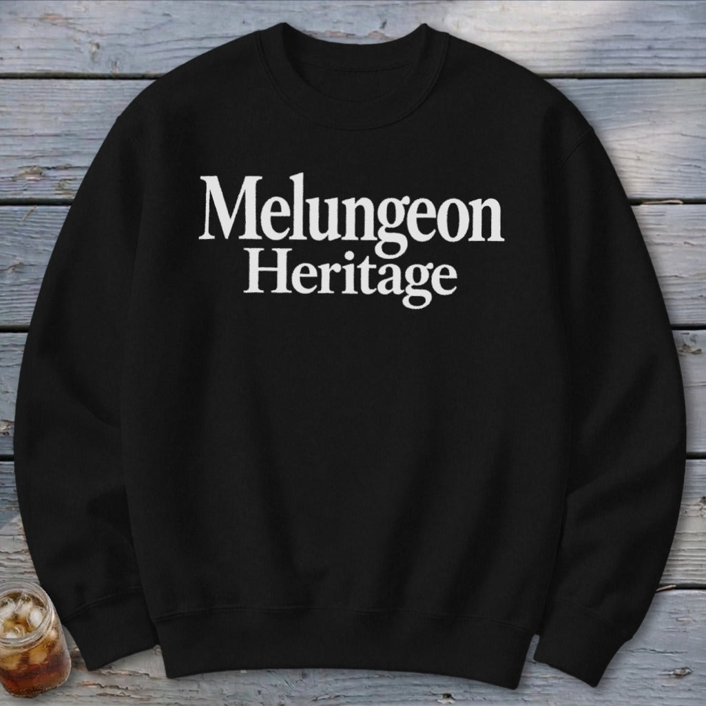 Black crewneck sweatshirt featuring white text that reads Melungeon Heritage centered on the chest, laid flat against a weathered wooden background.
