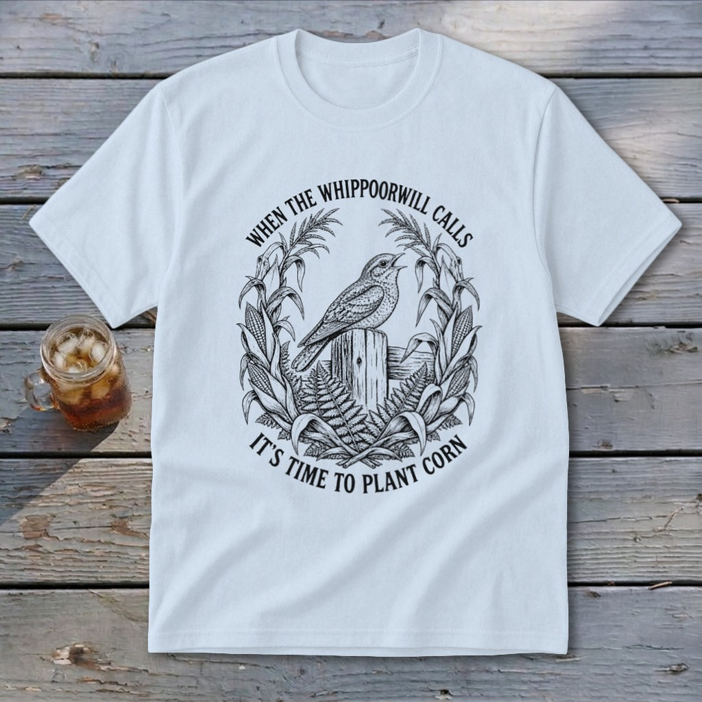 A white t-shirt featuring a black graphic of a bird perched on a wooden post, surrounded by a corn stalk wreath and the text When the Whippoorwill Calls, It's Time to Plant Corn.