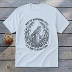 A white t-shirt featuring a black graphic of a bird perched on a wooden post, surrounded by a corn stalk wreath and the text When the Whippoorwill Calls, It's Time to Plant Corn.