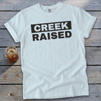 White t-shirt with "CREEK RAISED" printed in black text.