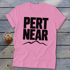 A pink crewneck t-shirt with rolled sleeves lies on a rustic wooden background, featuring black text that reads PERT NEAR above a mountain range graphic.