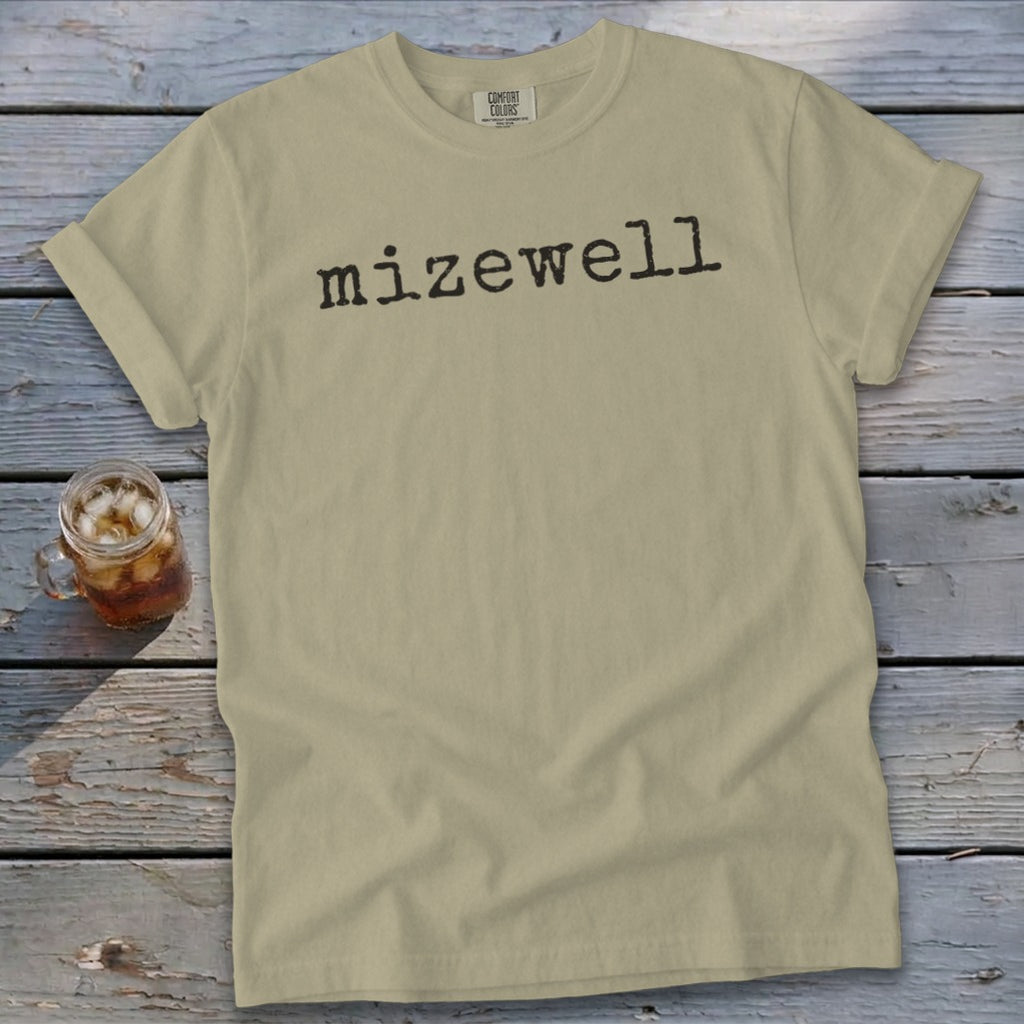 Khaki t-shirt with "mizewell" printed in black font, next to a glass of iced tea.