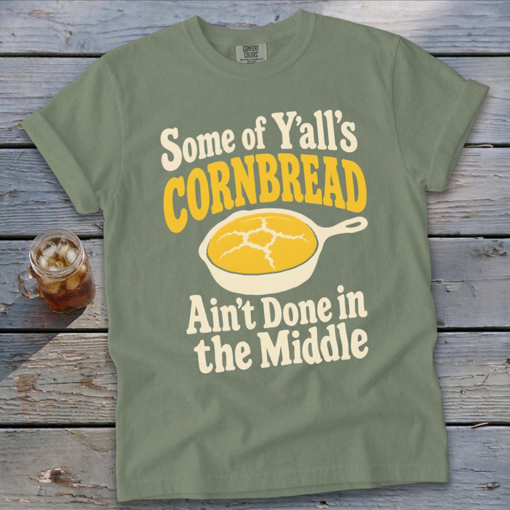 Sage green t-shirt featuring text and a skillet of cornbread laid flat on a rustic wooden background.
