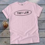 Light pink t-shirt with rolled sleeves featuring the text THEY LAW in a black oval design, laid on weathered wood.