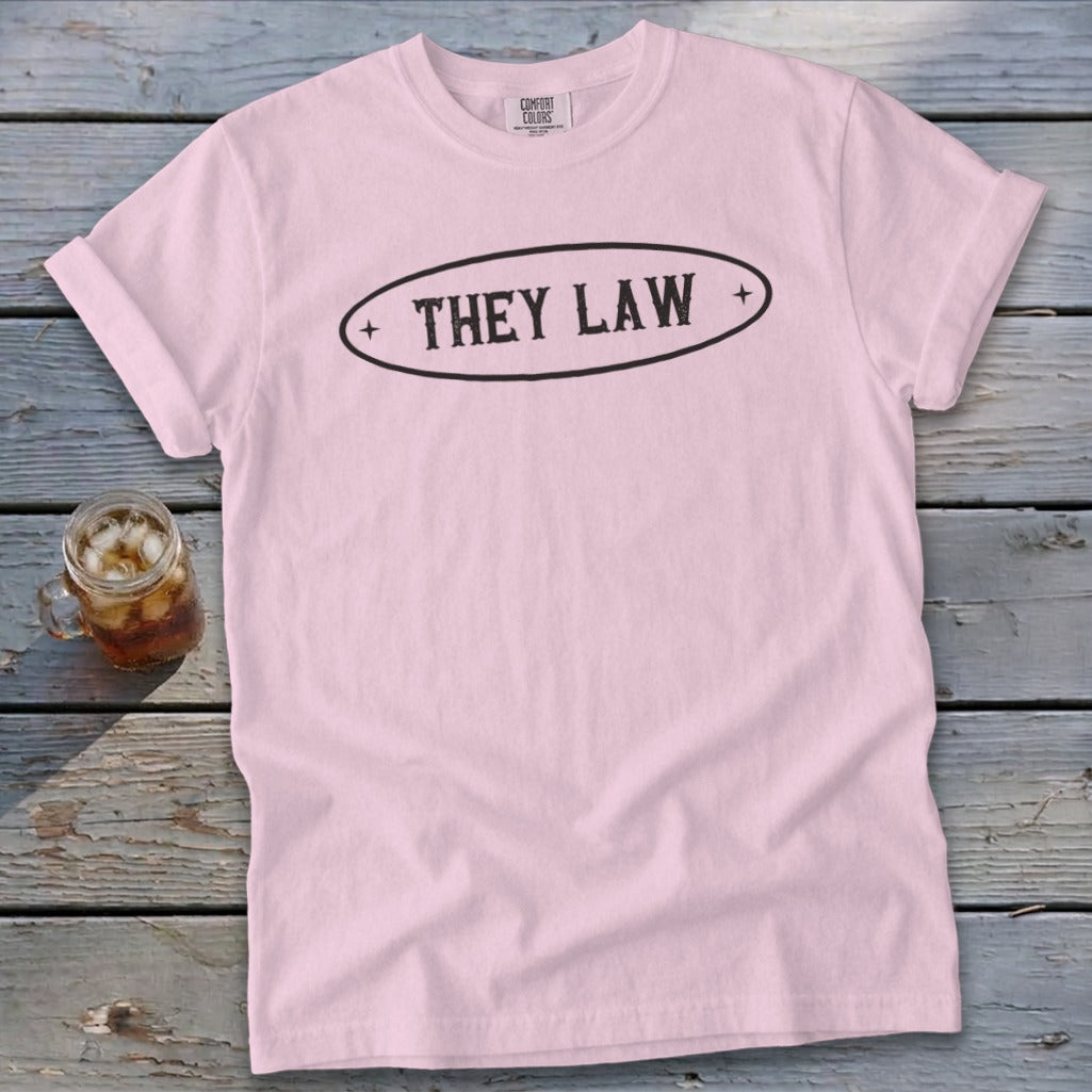 Light pink t-shirt with rolled sleeves featuring the text THEY LAW in a black oval design, laid on weathered wood.