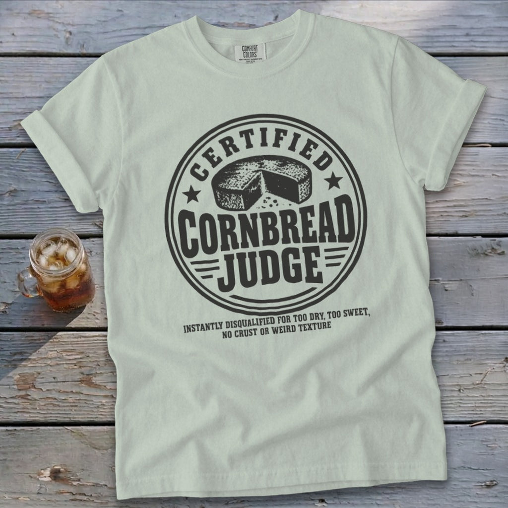 Light green t-shirt with a vintage round stamp graphic that reads Certified Cornbread Judge over a wooden background.