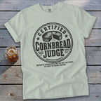 Light green t-shirt with a vintage round stamp graphic that reads Certified Cornbread Judge over a wooden background.