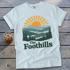 White t-shirt with a sunset over mountain layers and text "The Foothills".