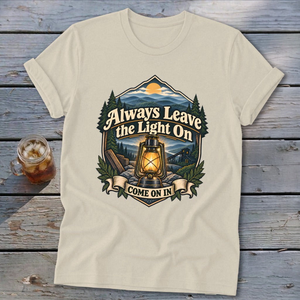 Cream t-shirt with graphic of lantern, mountains, trees, and text "Always Leave the Light On COME ON IN."