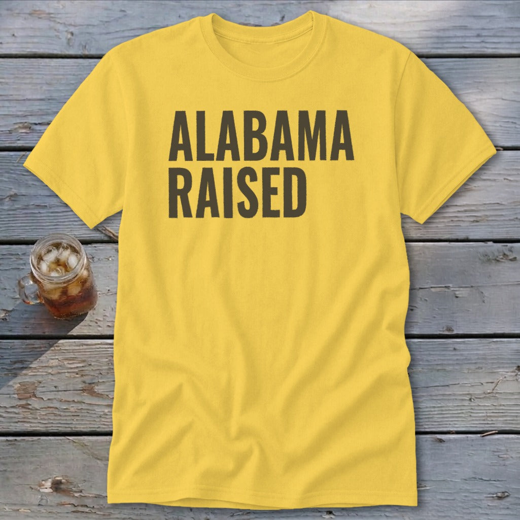 Yellow t-shirt with "ALABAMA RAISED" text on a wooden background.