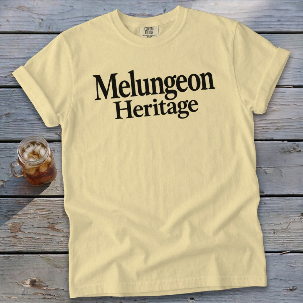 A light yellow t-shirt with black text reading Melungeon Heritage laid on a weathered wooden surface.