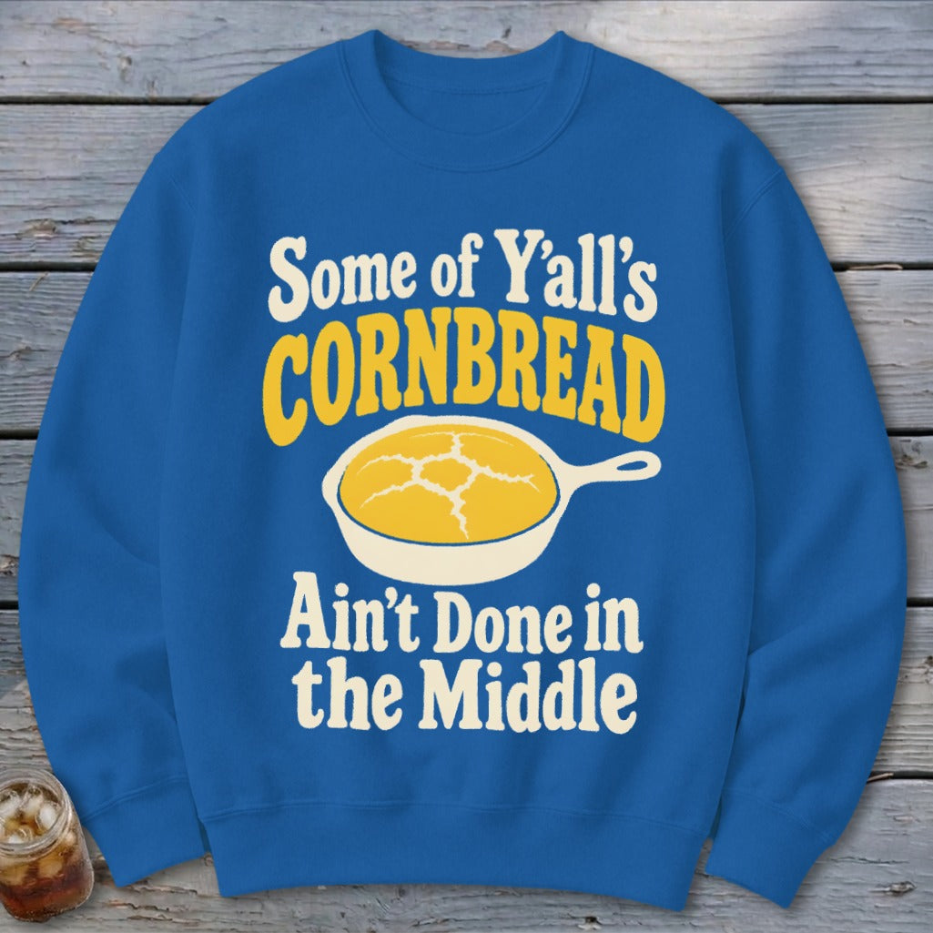 A blue crewneck sweatshirt featuring a graphic of cornbread in a skillet and text that reads Some of Y'all's Cornbread Ain't Done in the Middle.