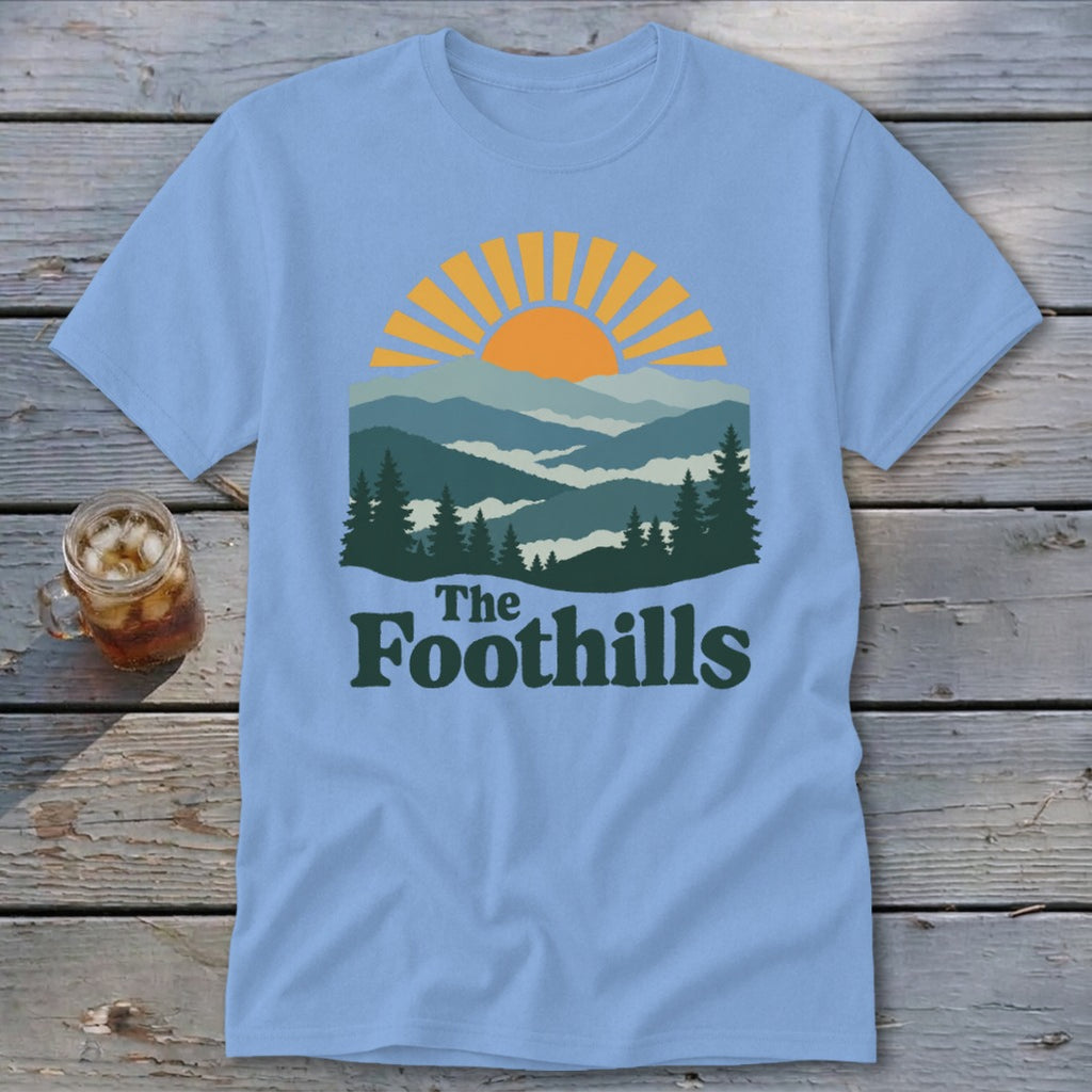 Light blue t-shirt with a mountain landscape and sunrise graphic, text reads "The Foothills".