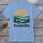 Light blue t-shirt with a mountain landscape and sunrise graphic, text reads "The Foothills".