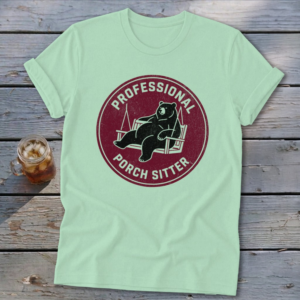 A mint green t-shirt with a red circular graphic showing a bear relaxing in a porch swing and the text "PROFESSIONAL PORCH SITTER".