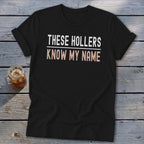 Black t-shirt with white text "THESE HOLLERS" and pink text "KNOW MY NAME."