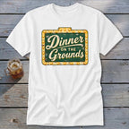 A white t-shirt featuring a green and gold sign graphic that reads Dinner on the Grounds.