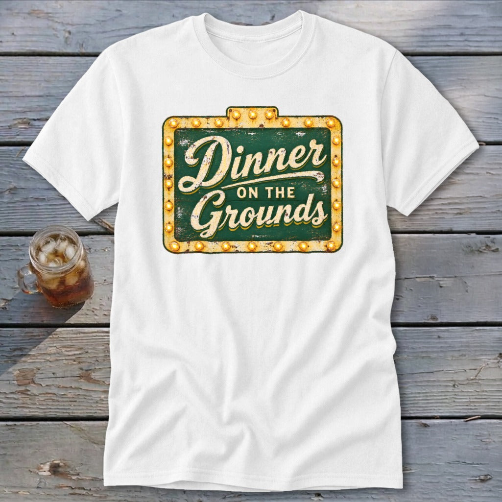 A white t-shirt featuring a green and gold sign graphic that reads Dinner on the Grounds.