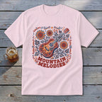 A light pink t-shirt with a vintage-style graphic of an acoustic guitar, sunflowers, and the text MOUNTAIN MELODIES.