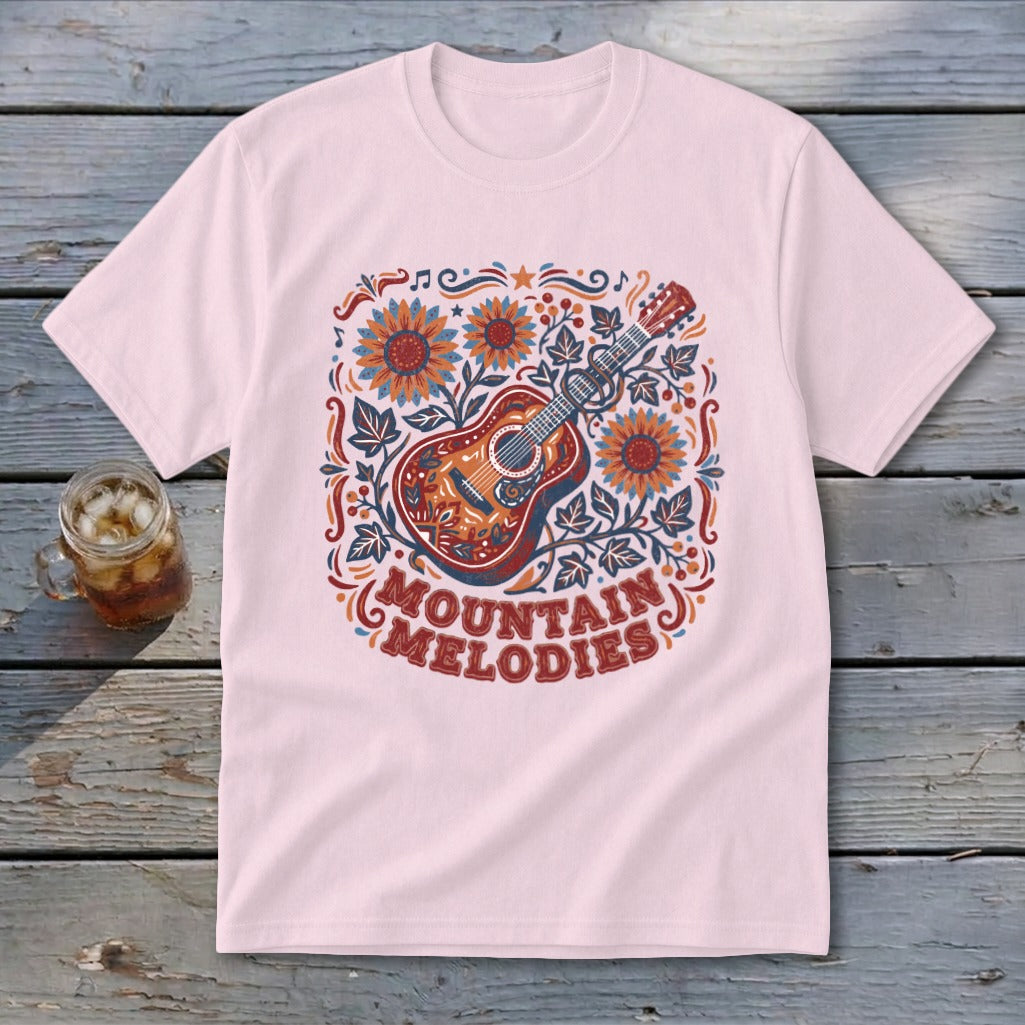 A light pink t-shirt with a vintage-style graphic of an acoustic guitar, sunflowers, and the text MOUNTAIN MELODIES.