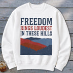 A white crewneck sweatshirt featuring blue and red mountain graphics and the text Freedom Rings Loudest in These Hills.