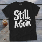 A black t-shirt with white handwritten text reading Still A-Goin' on a wooden background.