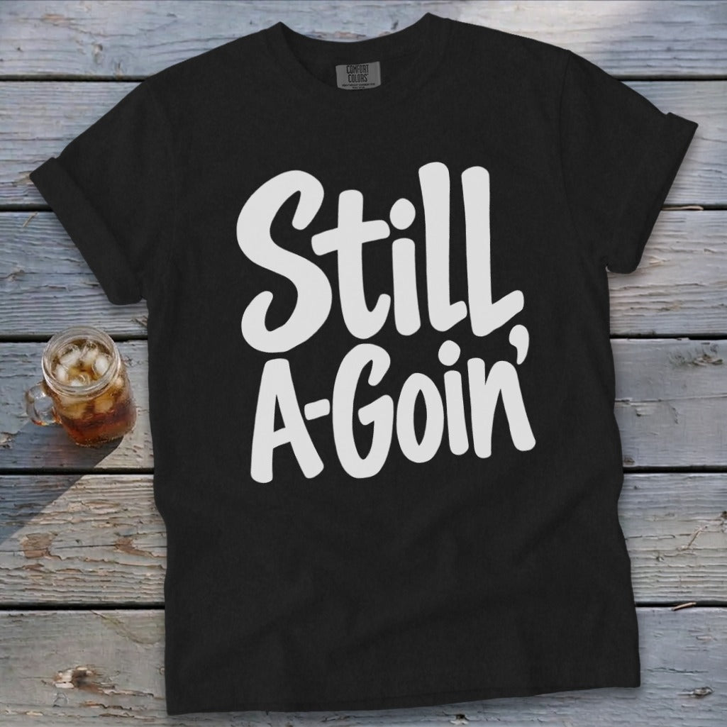 A black t-shirt with white handwritten text reading Still A-Goin' on a wooden background.