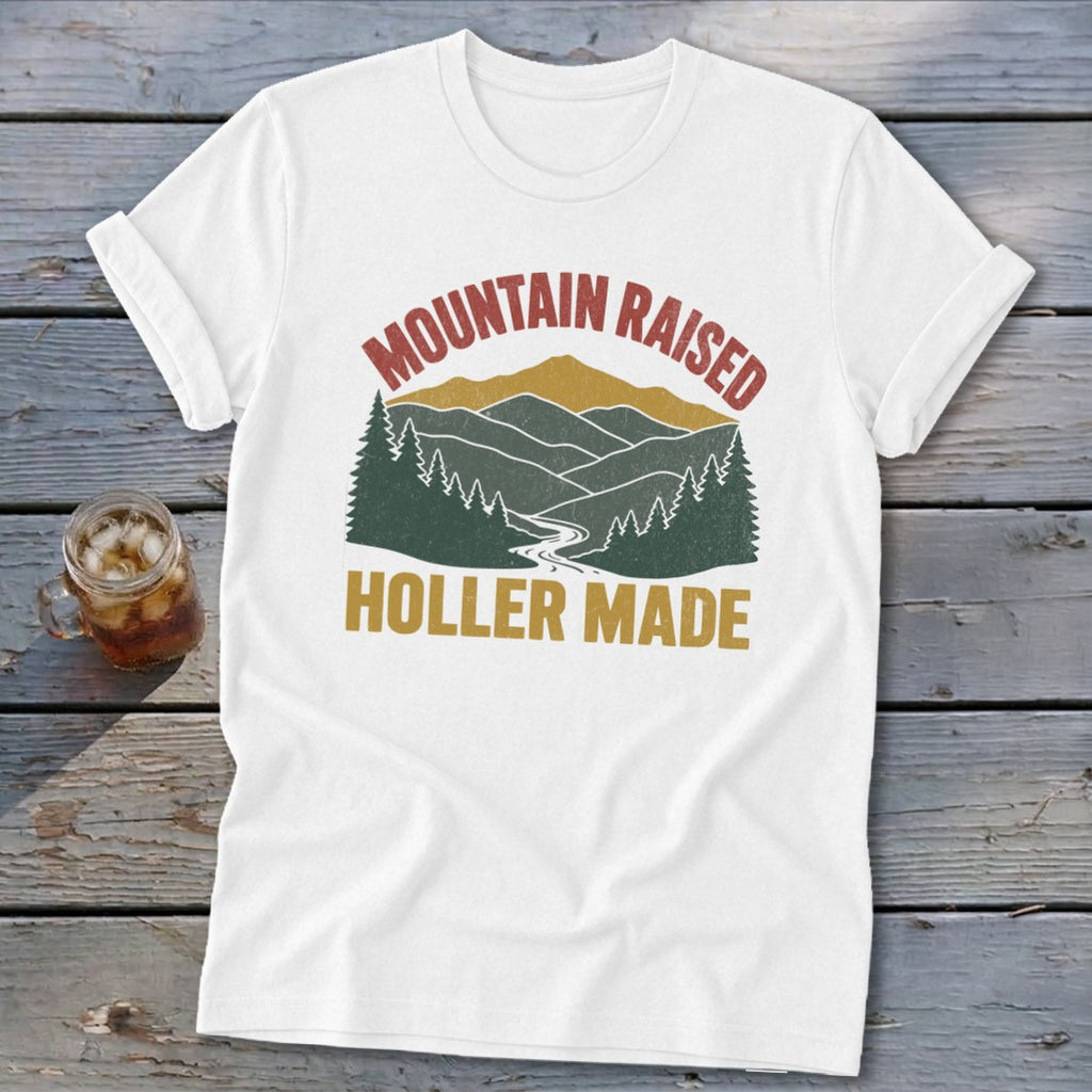 A white short-sleeved t-shirt featuring a retro graphic of mountains, trees, and a river with text reading "MOUNTAIN RAISED HOLLER MADE".