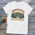 A white short-sleeved t-shirt featuring a retro graphic of mountains, trees, and a river with text reading "MOUNTAIN RAISED HOLLER MADE".