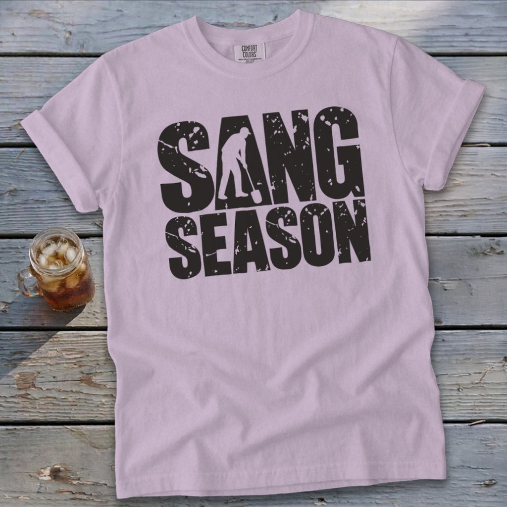 A light purple t-shirt featuring distressed black text reading SANG SEASON, lying on a wooden surface.