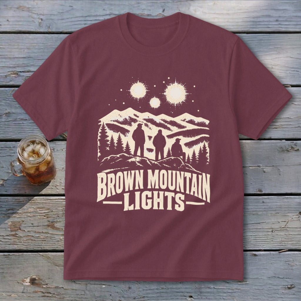 Maroon t-shirt featuring a cream graphic of mountains, three silhouetted figures, glowing lights, and text reading Brown Mountain Lights.