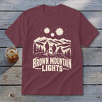 Maroon t-shirt featuring a cream graphic of mountains, three silhouetted figures, glowing lights, and text reading Brown Mountain Lights.