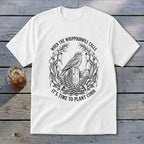 A white t-shirt featuring a black graphic of a bird on a post, surrounded by corn stalks and ferns, with the text "WHEN THE WHIPPOORWILL CALLS IT'S TIME TO PLANT CORN."