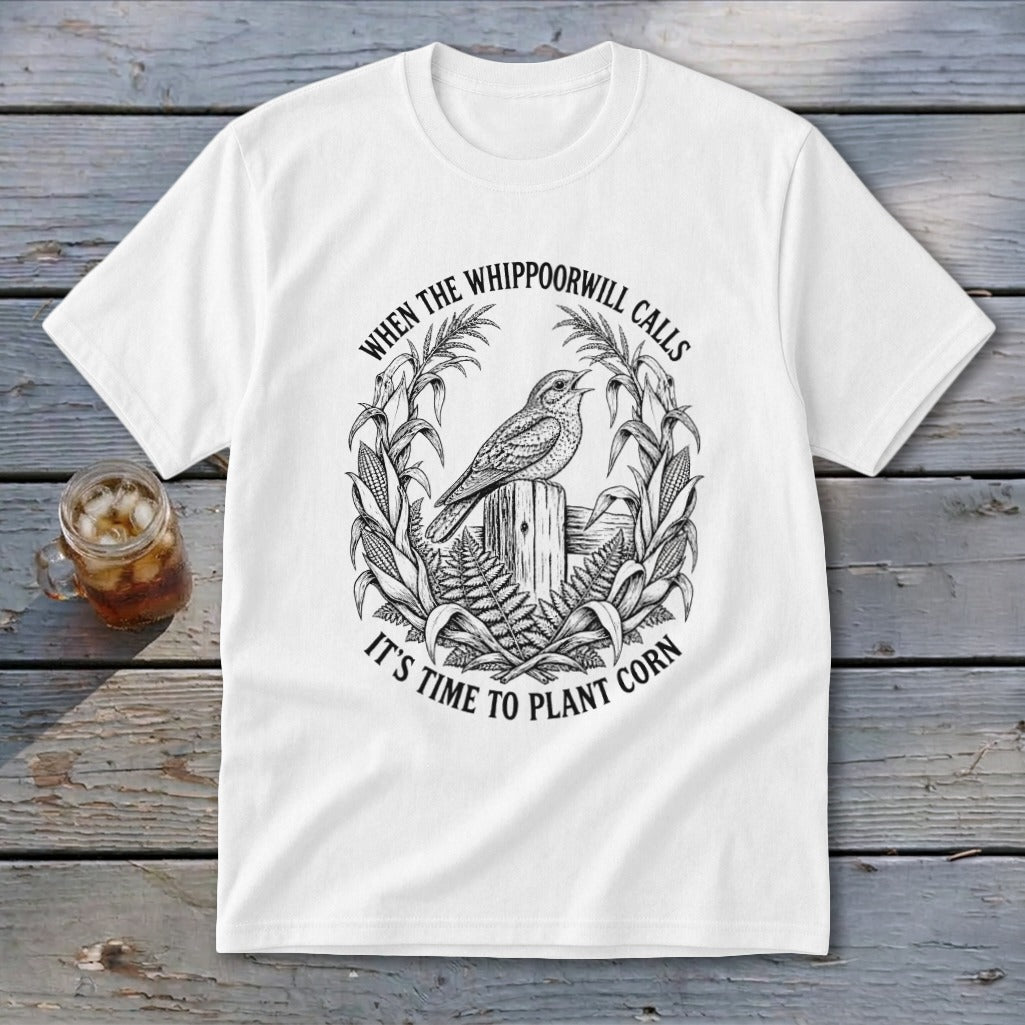 A white t-shirt featuring a black graphic of a bird on a post, surrounded by corn stalks and ferns, with the text "WHEN THE WHIPPOORWILL CALLS IT'S TIME TO PLANT CORN."