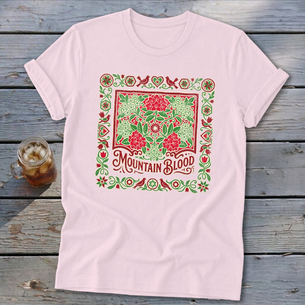 Pink t-shirt with a decorative square design featuring red flowers, green foliage, and "Mountain Blood" text.