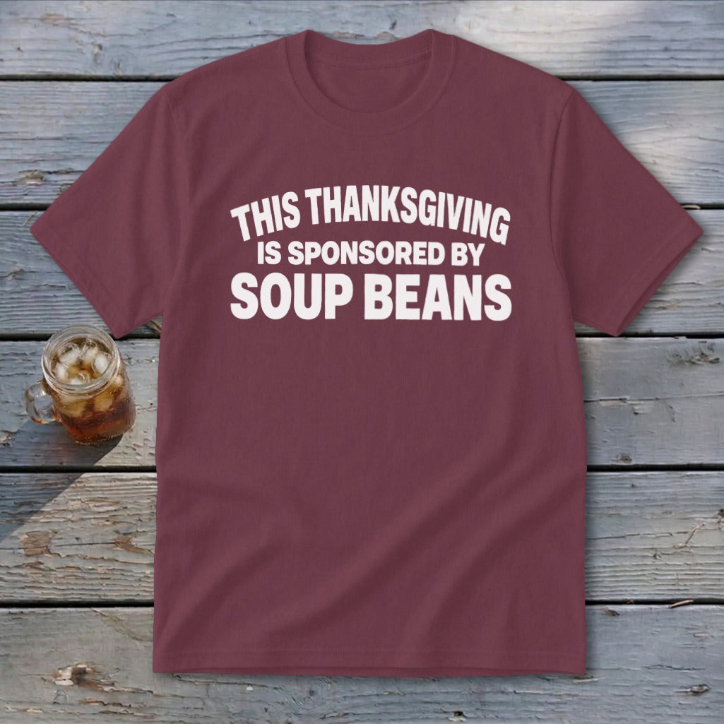 A maroon t-shirt with white text reading THIS THANKSGIVING IS SPONSORED BY SOUP BEANS laid on a rustic wooden table next to a glass of iced tea.
