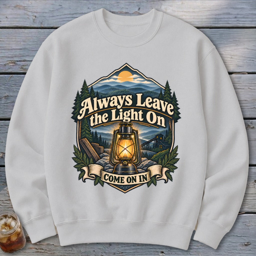 White sweatshirt with a graphic of a lantern, mountains, and text "Always Leave the Light On COME ON IN".