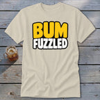 A cream-colored t-shirt rests on a wooden surface, featuring the black-bordered text BUM FUZZLED in yellow and white.