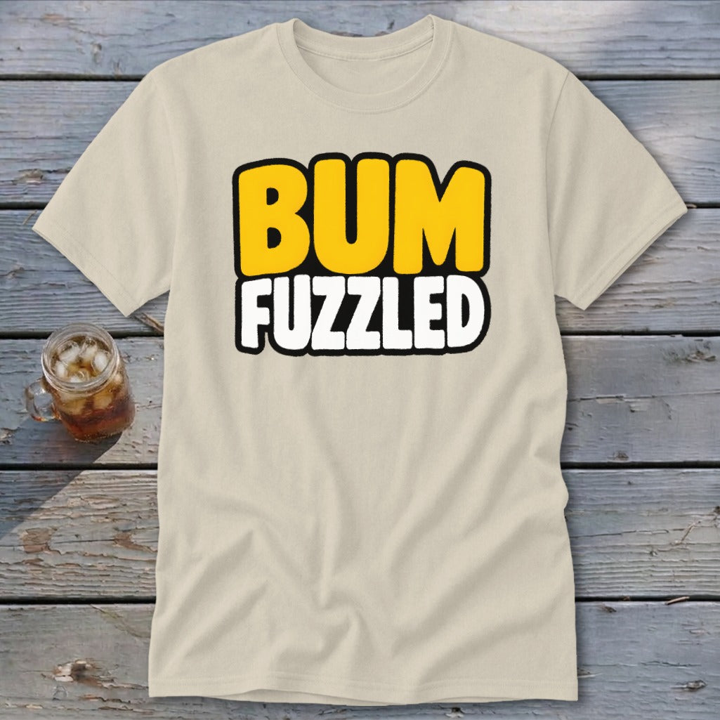 A cream-colored t-shirt rests on a wooden surface, featuring the black-bordered text BUM FUZZLED in yellow and white.