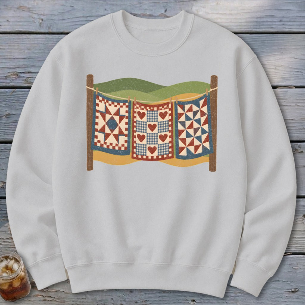 Light grey crewneck sweatshirt featuring a graphic of three patchwork quilts hanging on a clothesline against hills.