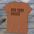 Orange t-shirt with "NEW YORK RAISED" text and a drink on wooden planks.